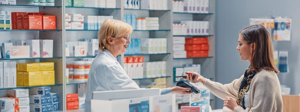 Apothekerin übergibt ein Produkt an eine Kundin an der Theke einer Apotheke mit vielen Medikamenten im Hintergrund.